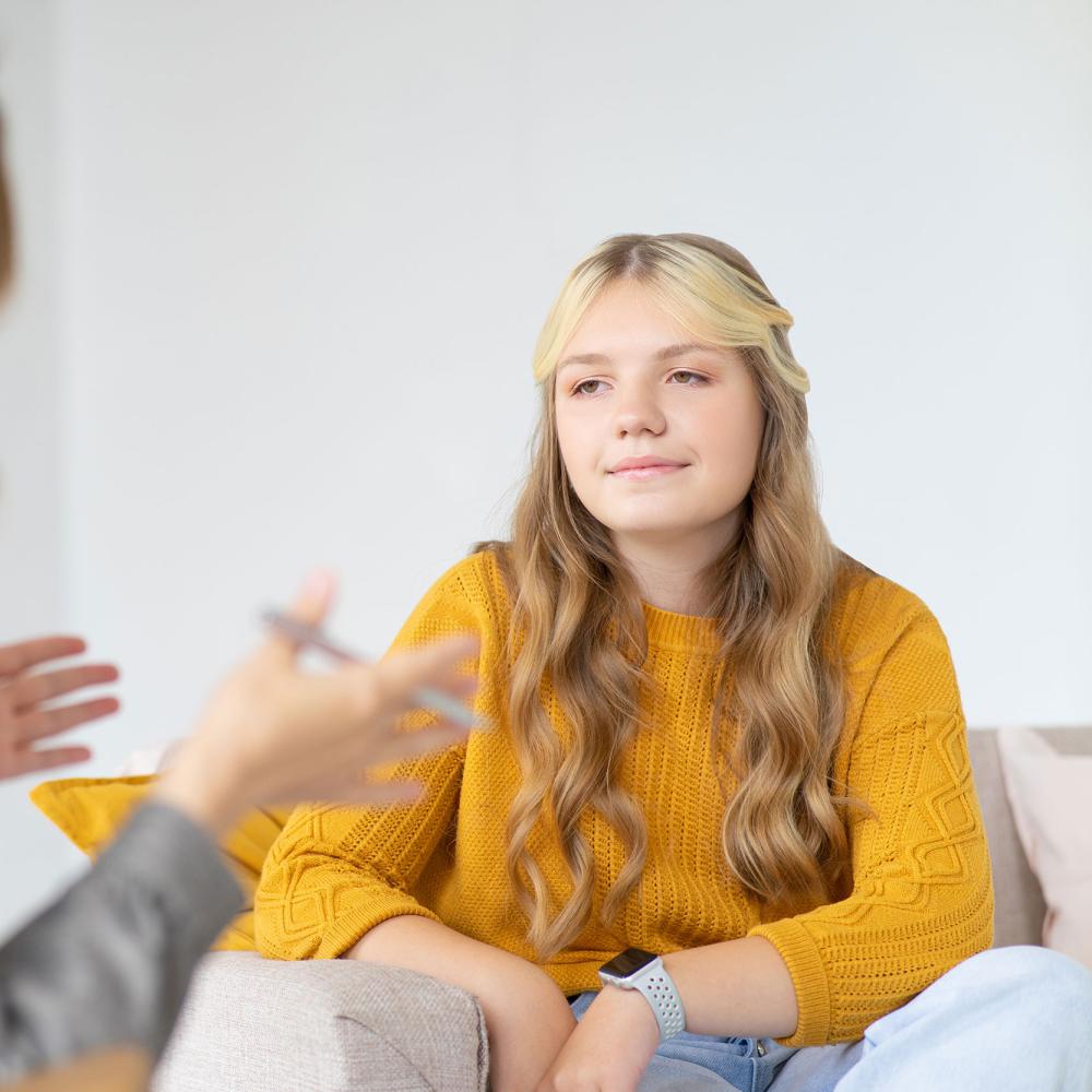 A girl sits and listens to a woman gesturing with her hands and holding a pen