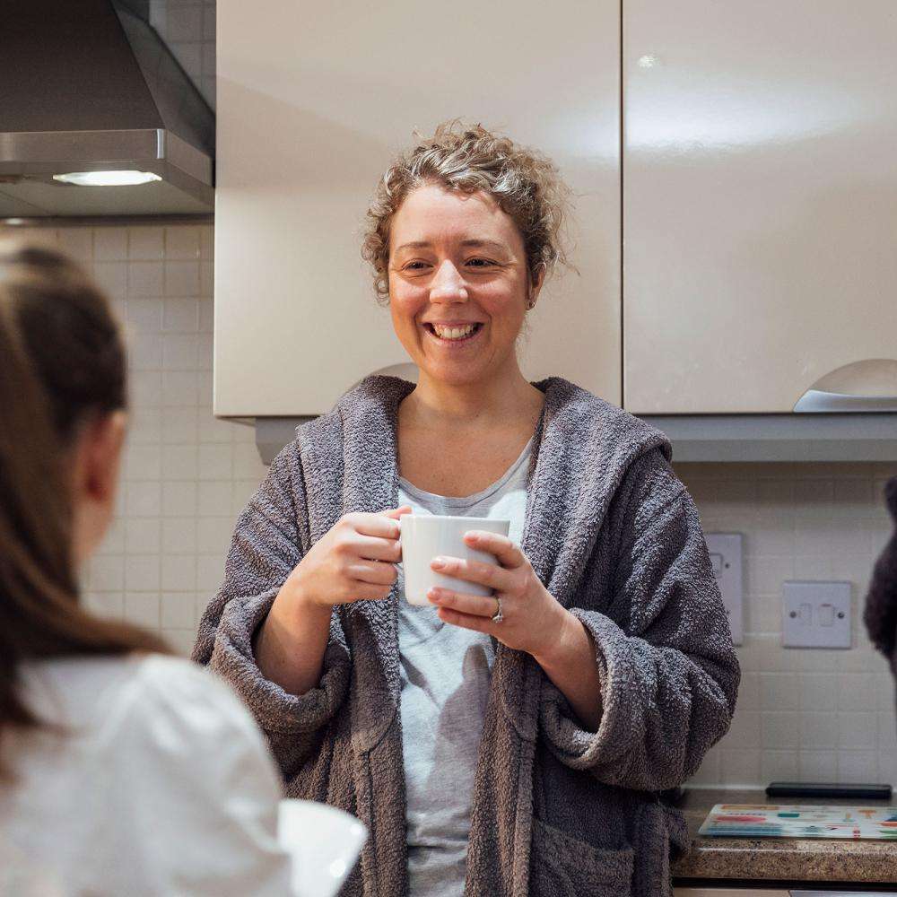 A woman in a dressing gown smiles while holding a mug. She is with a boy and a girl