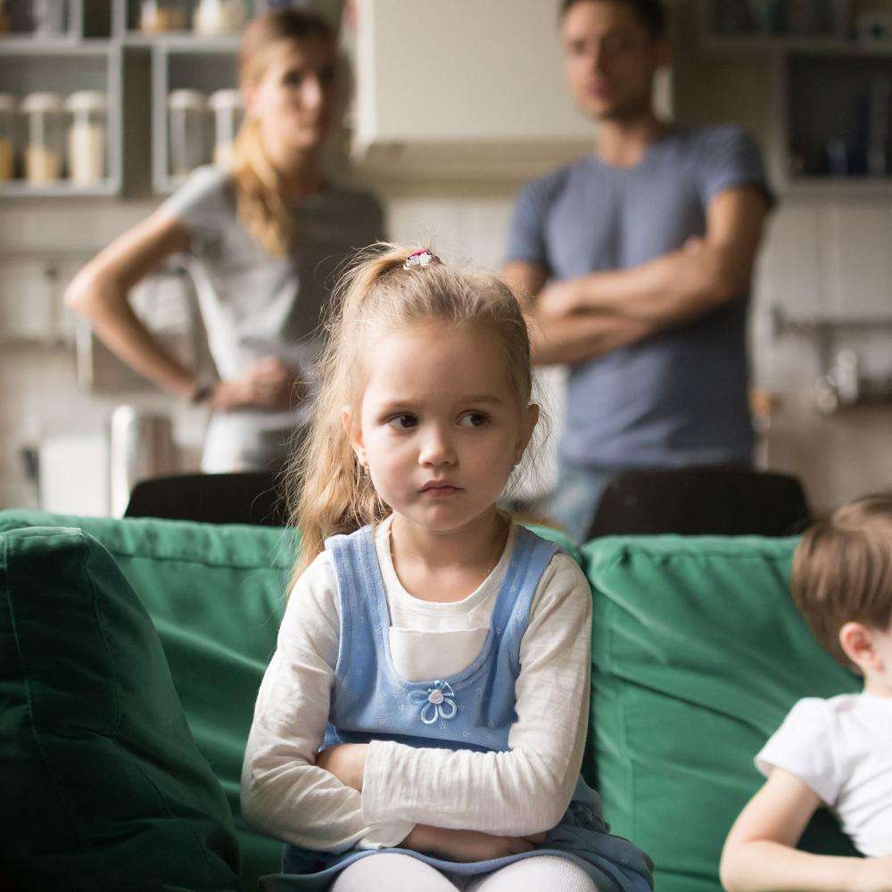 Two children look sad sat on sofa while parents stand in background