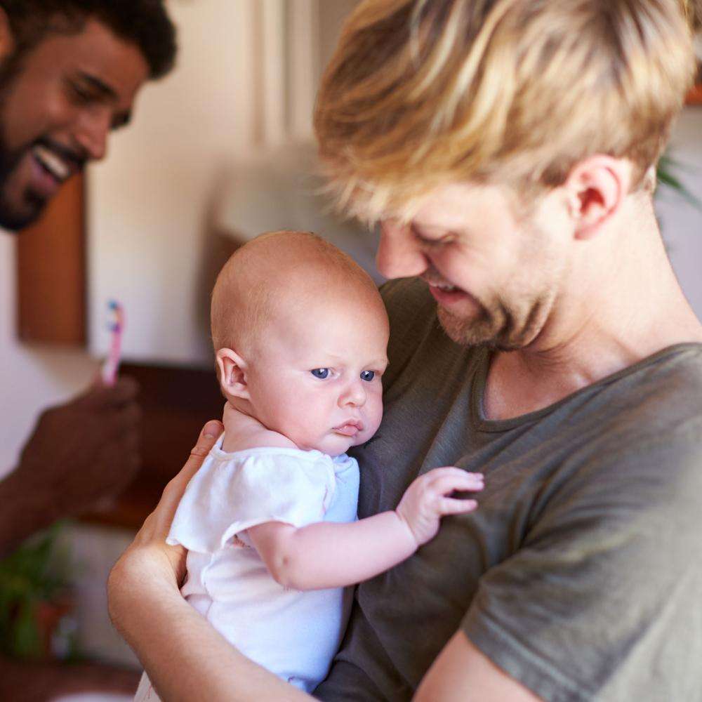 A man smiles and holds a baby. His male partner laughs while brushing his teeth