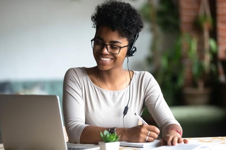 A woman working at a computer with a headset on smiles while writing notes