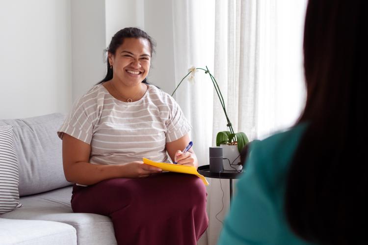 A woman sits and smiles while holding a pad and pen